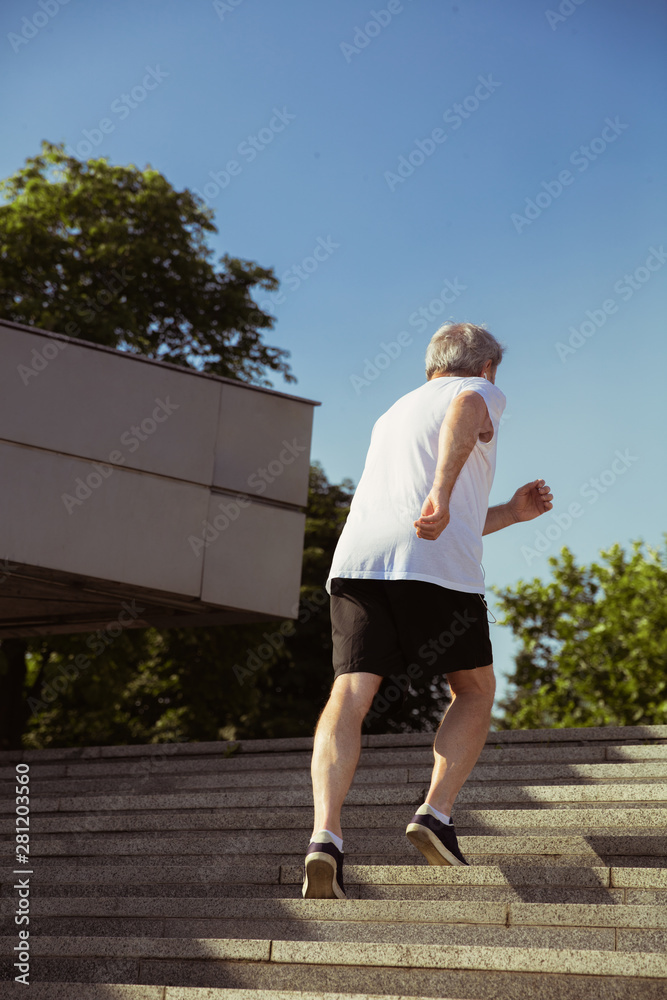 Senior man as runner with armband or fitness tracker at the city's street. Caucasian male model practicing jogging and cardio trainings in summer's morning. Healthy lifestyle, sport, activity concept.