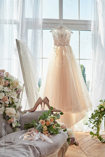 Beautiful beige women's shoes and bridal bouquet on chesterfield sofa with wedding dress in blur hanging on hanger against window in hotel room, copy space