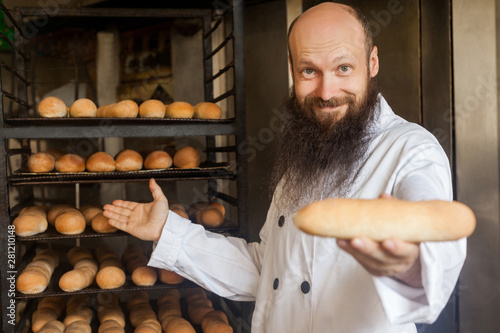 Please take it. Portrait of attractive positive young adult baker with long beard in white uniform standing in his workplace and giving you fresh loaf, Indoor, profession concept, looking at camera