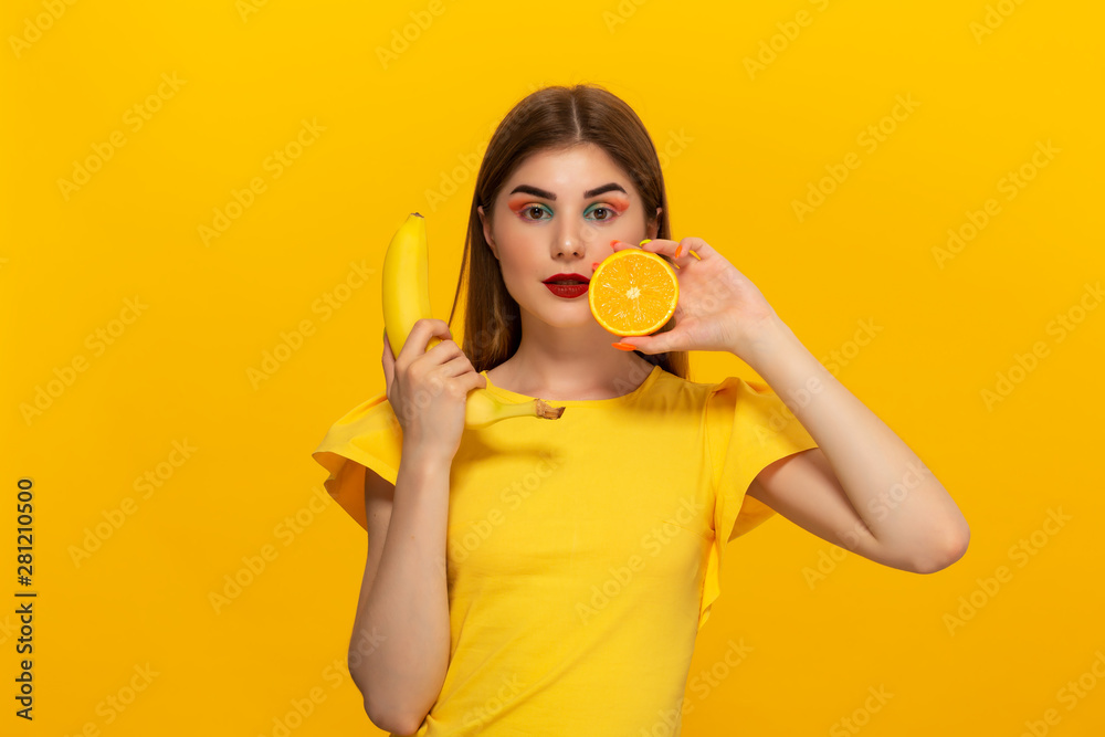Young model in a yellow tee holds banana and orange standing over yellow background. Young model in a yellow tee holds banana and orange standing over yellow background.