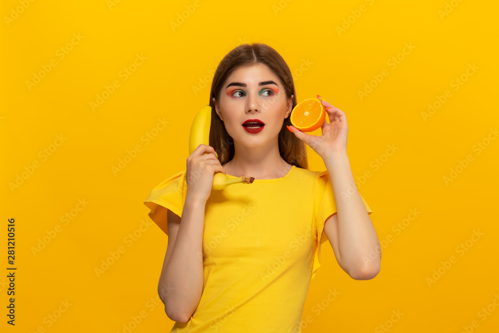 Young model in a yellow tee holds banana and orange standing over yellow background. Young model in a yellow tee holds banana and orange standing over yellow background.