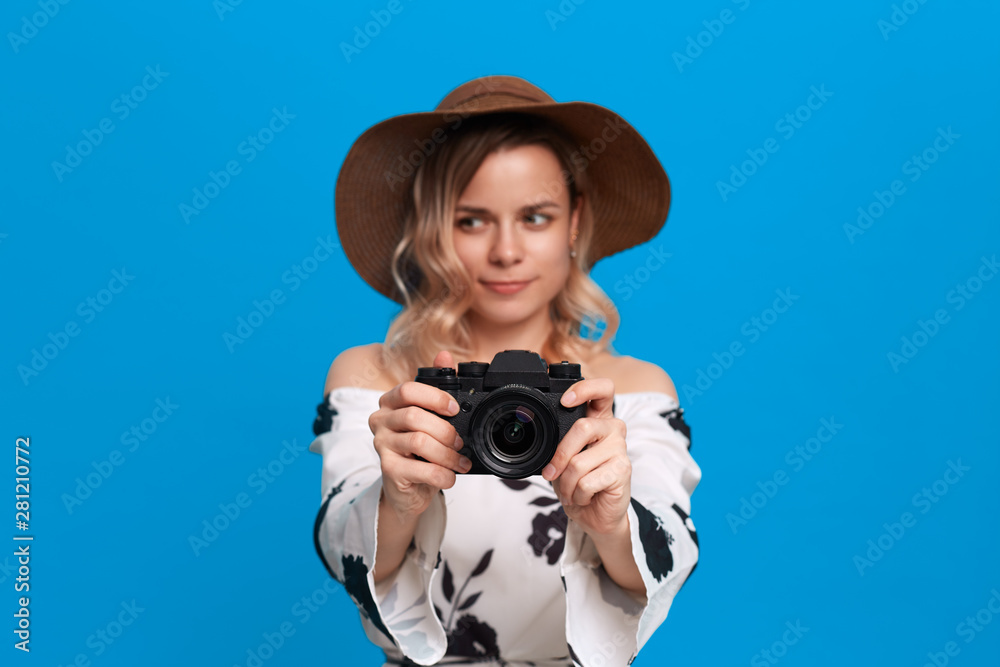 Portrait of a model with curly blond hair in a sundown hat and white dress takes photos with retro camera standing on a blue background. Portrait of a model with curly blond hair in a sundown hat and white dress takes photos with retro camera standing on a blue background.