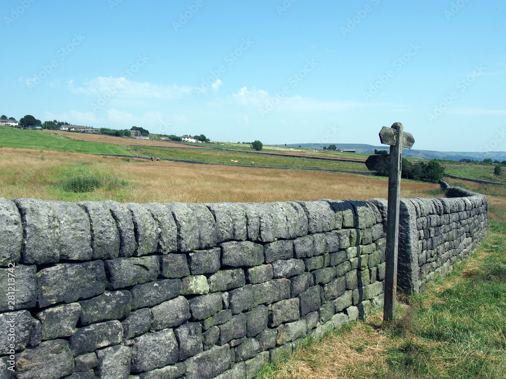 rural scene with a long stone wall and wooden signpost next to a gate in hillside meadows with farmhouses and hills in the distance in blackshaw head in west yorkshire