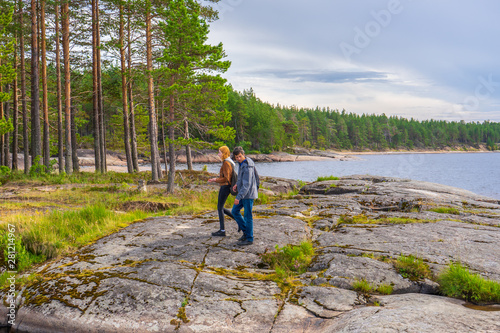 Middle aged man and young lady walking on northern lake shore in summer day. Tourists against picturesque landscape. Travelling and discovering distant places of Earth. Onega lake, Karelia, Russia