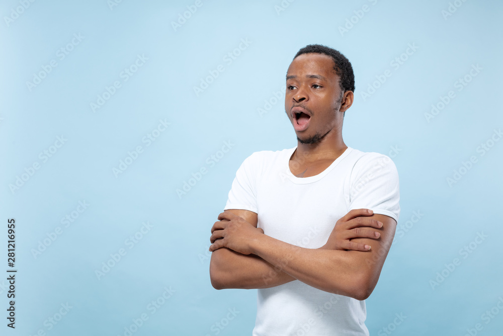 Half-length close up portrait of young african-american man in white shirt on blue background. Human emotions, facial expression, ad, sales, concept. Standing hands crossed, shocked and astonished. Half-length close up portrait of young african-american man in white shirt on blue background. Human emotions, facial expression, ad, sales, concept. Standing hands crossed, shocked and astonished.