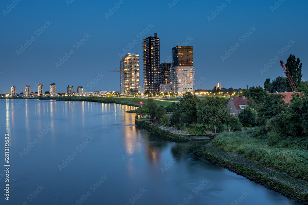 luminated tall apartment buildings at the riverside after sunset. Spijkenisse skyline at the Oude Maas river. With small house surrounded by green and wave breakers rocks at the shoreline