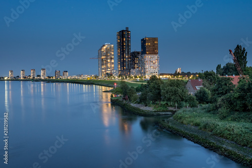 luminated tall apartment buildings at the riverside after sunset. Spijkenisse skyline at the Oude Maas river. With small house surrounded by green and wave breakers rocks at the shoreline © Frank