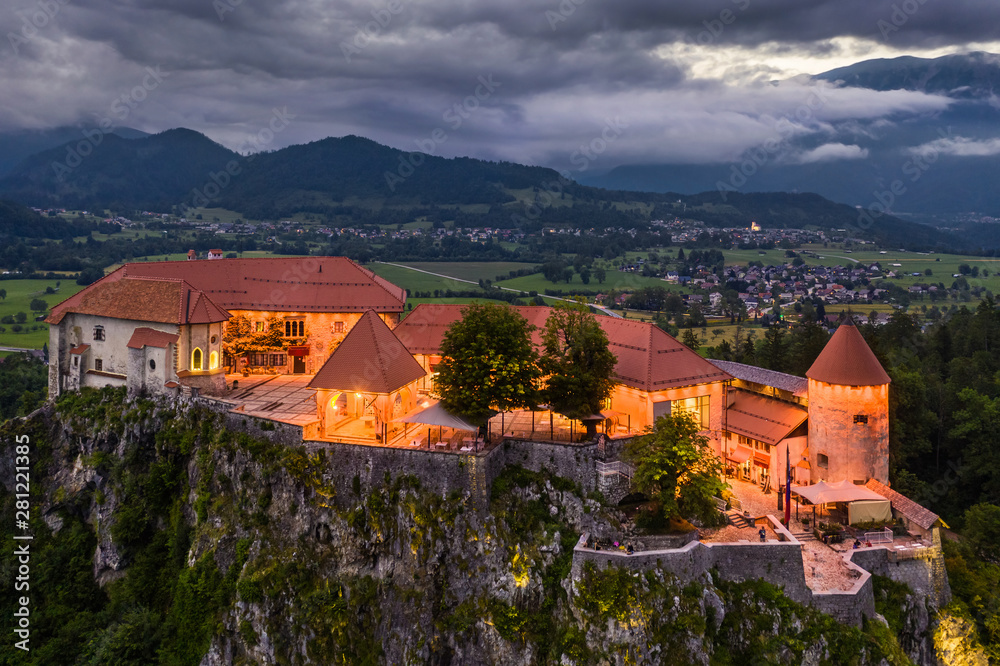 Bled, Slovenia - Aerial view of beautiful illuminated Bled Castle (Blejski Grad) with the Julian Alps at background at blue hour with dramatic sky