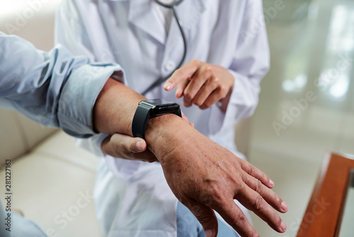 Close-up of senior patient with wristwatch on his hand sitting with doctor who pointing at wristwatch and checking his pulse