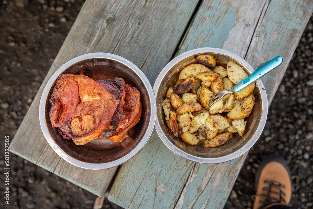 Fototapeta premium Metal bowls with lamb steaks and potatoes on wooden picnic table. Top view.
