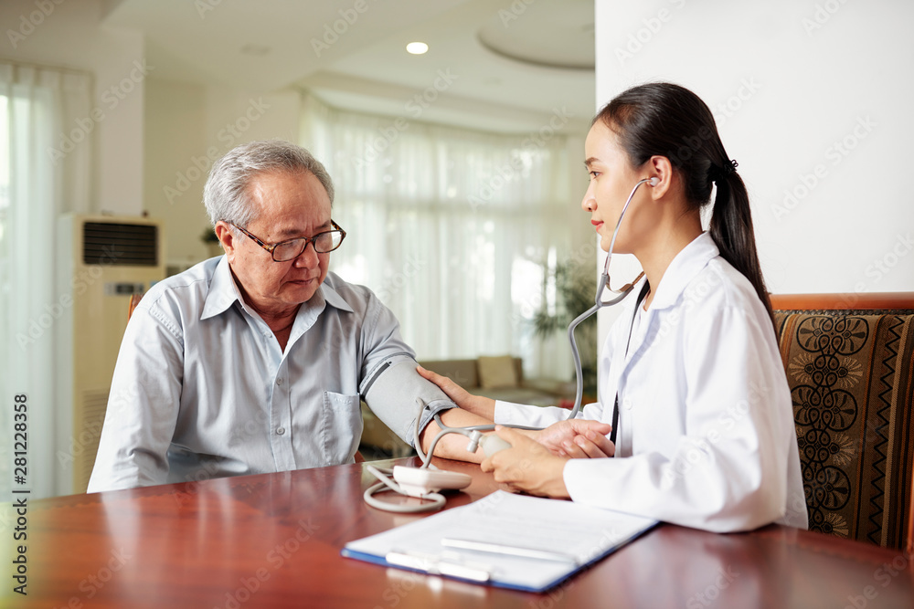 Asian young nurse sitting at the table together with elderly man and examining his blood pressure during her visit at home Asian young nurse sitting at the table together with elderly man and examining his blood pressure during her visit at home