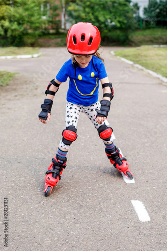 A small child in bright clothes, in a red helmet and protection goes on the road on rollers. Children's sport, active leisure.