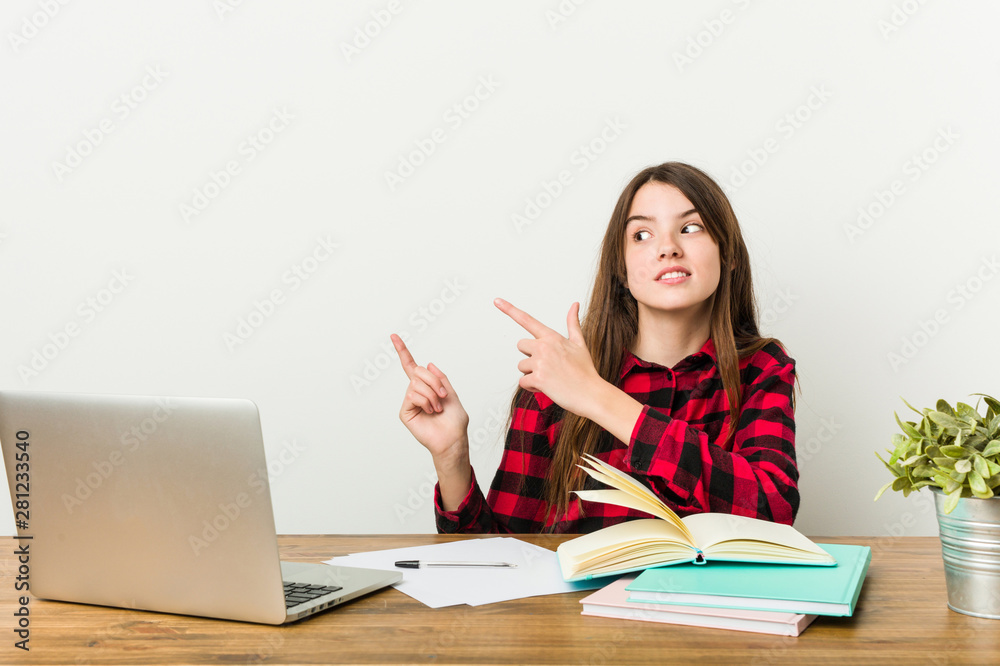 Young teenager going back to her routine doing homeworks shocked pointing with index fingers to a copy space. Young teenager going back to her routine doing homeworks shocked pointing with index fingers to a copy space.
