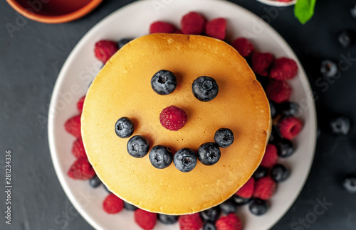 American pancakes with raspberries, fresh blueberries and honey. Healthy breakfast on concrete background, funny face on pancakes painted with blueberries