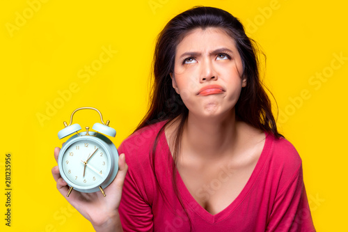Beautiful young woman hold alarm clock and gets lazy, boring, when she has to wake up early for work on Monday morning. Pretty girl doesn’t want to work because she is lazy lady. She make frown face