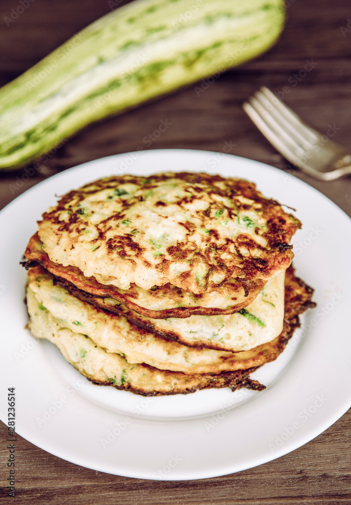 Fried zucchini pancakes stacked on plate, healthy organic summer food made with zucchini. Brown wood board background.
