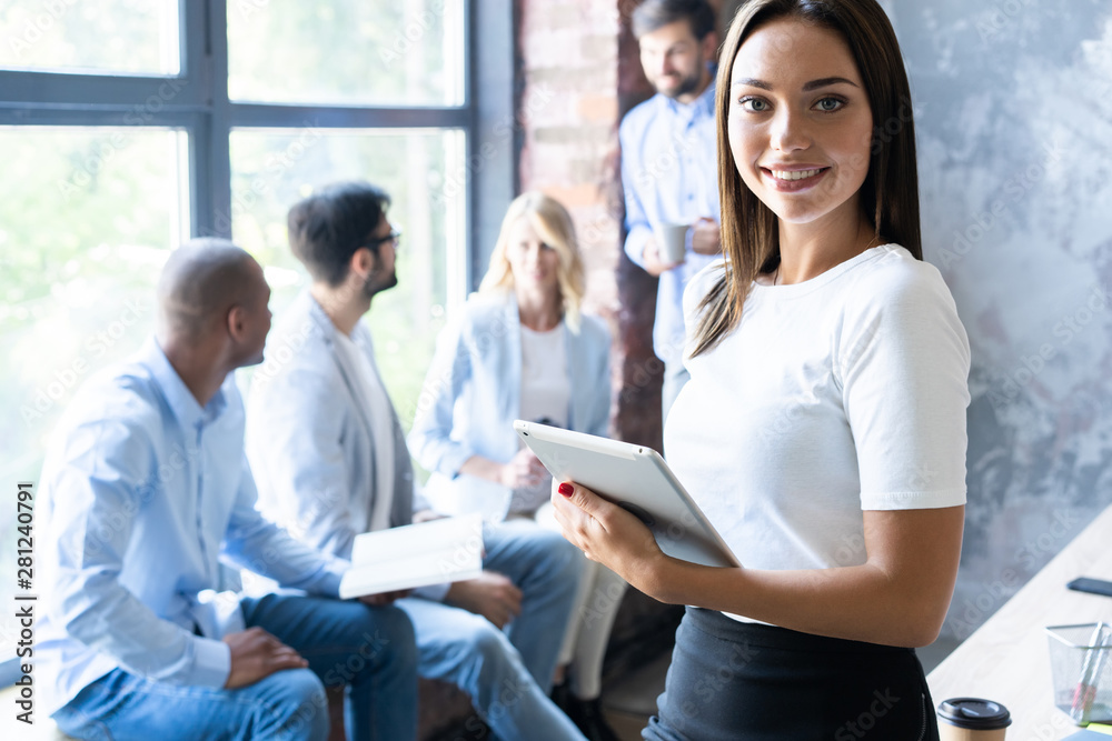 Beautiful young business lady is looking at camera and smiling while her colleagues are working in the background. Beautiful young business lady is looking at camera and smiling while her colleagues are working in the background.