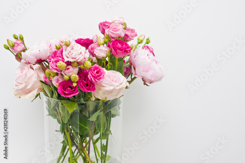Bouquet of pink roses, peony flowers in glass vase on a shelf against white wall background.
