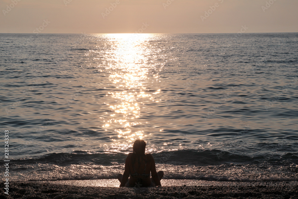 Girl sitting on sandy beach next to sea and enjoys watching sunset, reflection of sun rays over sea. Silhouette of woman by ocean. Light and shadow, woman rests during sunset at tropical landscape.