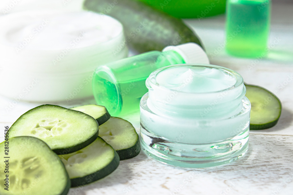 Face cream in a glass jar with cucumber extract next to slices of fresh cucumbers on a white wooden background. Face cream in a glass jar with cucumber extract next to slices of fresh cucumbers on a white wooden background.
