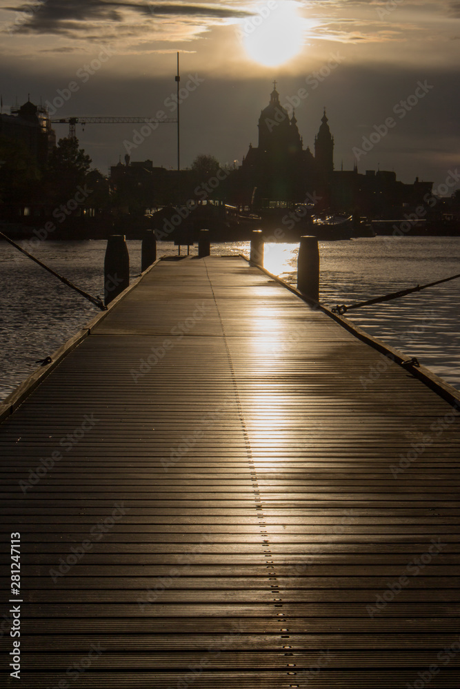 Obraz premium Wooden pier with boats at sunset in Amsterdam dock. Amsterdam city silhouettes in evening dusk light. Cathedral silhouette on evening sky in Netherlands. Downtown in Amsterdam dock. 