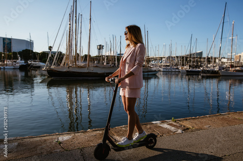 Young beautiful woman in a pink suit riding an electric scooter in the port of the sea,  modern girl, new generation, electric transport, ecology, ecological transport, dawn, electric skateboard