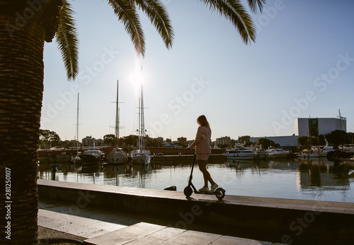 Young beautiful woman in a pink suit riding an electric scooter in the port of the sea, modern girl, new generation, electric transport, ecology, ecological transport, dawn, electric skateboard