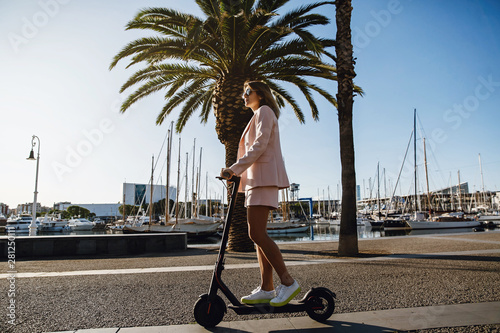 Young beautiful woman in a pink suit riding an electric scooter in the port of the sea,  modern girl, new generation, electric transport, ecology, ecological transport, dawn, electric skateboard