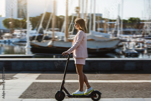 Young beautiful woman in a pink suit riding an electric scooter in the port of the sea, modern girl, new generation, electric transport, ecology, ecological transport, dawn, electric skateboard