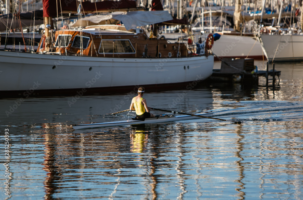 Obraz premium A man in a yellow shirt kayaking in a port near yachts and boats at dawn. Summer travel kayaking. Man paddling canoe. Exploring sea on vacation.