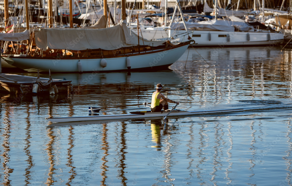 A man in a yellow shirt kayaking in a port near yachts and boats at dawn. Summer travel kayaking. Man paddling canoe. Exploring sea on vacation.