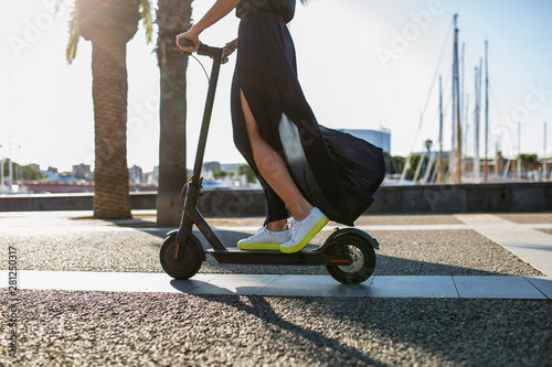 Young beautiful woman in a dark dress riding an electric scooter to work, female legs on electric scooter, electric transport, ecology, ecological transport, sunset, electric skateboard, 