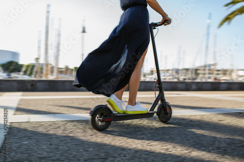 Young beautiful woman in a dark dress riding an electric scooter to work, female legs on electric scooter, electric transport, ecology, ecological transport, sunset, electric skateboard, 