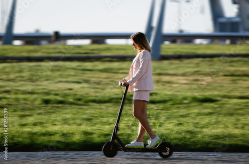 Young beautiful woman in a pink suit riding an electric scooter in the port of the sea, modern girl, new generation, electric transport, ecology, ecological transport, dawn, electric skateboard