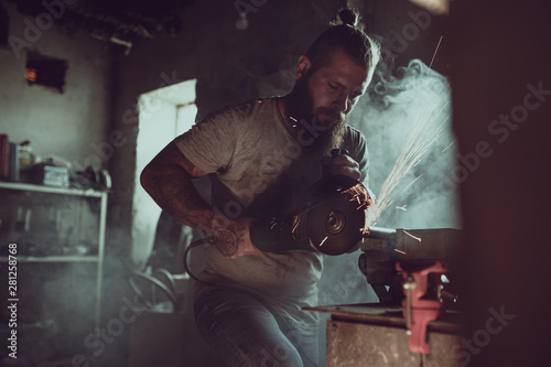 Handsome brutal male with a beard repairing a motorcycle in his garage working with a circular saw. In the garage a lot of sparks and smoke from sawing