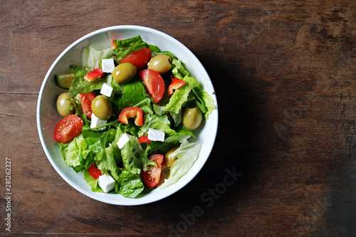 Traditional greek salad with fresh ingredients, feta cheese, olives, red tomatoes, cucumbers and greens in white ceramic bowl on wood textured background. Top view, close up, copy space.