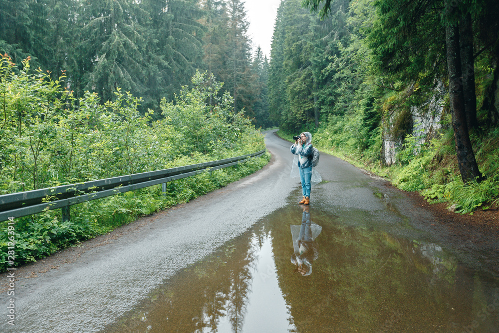 traveller girl  in the rain takes a picture with t camera on the mountainside in the forest. Young woman in raincoat  traveling with backpack and photo camera in beautiful evergreen forest