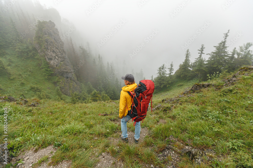 Fototapeta premium Traveler with backpack and mountain panorama. Man with big backpack and trekking poles standing on the edge and watching to the distant mountain range
