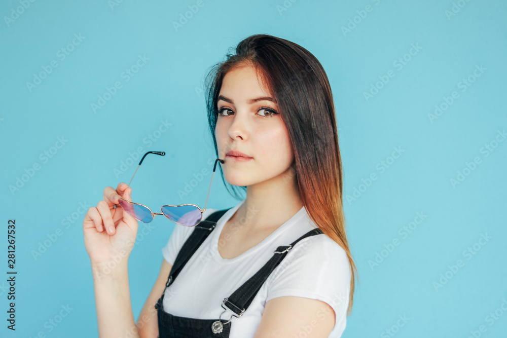 Beautiful girl in heart shape sunglasses with dark long hair in white t-shirt isolated on blue background Beautiful girl in heart shape sunglasses with dark long hair in white t-shirt isolated on blue background