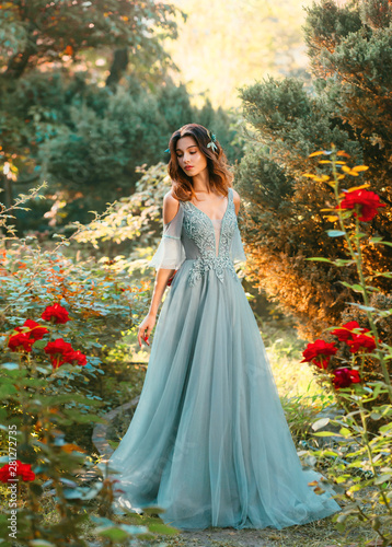 Affectionate bride in light blue dress in green garden with red lush roses, princess with dark hair looks down, pretty model poses for charming photo with creative colors, symbol of passing summer © kharchenkoirina