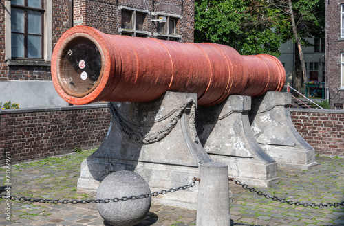 Gent, Flanders, Belgium -  June 21, 2019: Historic large, red, Dulle Griet Cannon with stone cannon ball. Brick wall with window and green foliage as background. photo