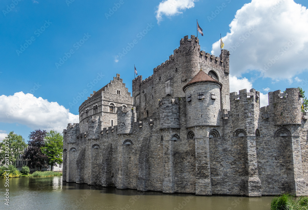 Gent, Flanders, Belgium -  June 21, 2019: Gray stone cast;e and ramparts of Gravensteen, historic medieval castle of city, behind its moad against blue sky with white clouds. Flags on top, green folia