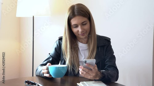 Beautiful Woman in a leaser jacket using a smart phone and holding cap while sitting at the table indoors photo