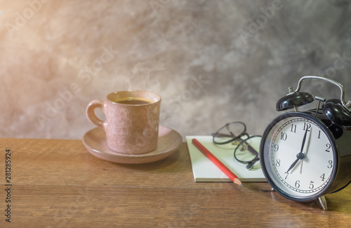 Close-up of retro alarm black clock vintage style with Blank open book, red pencil and glasses cup coffee pastel pink old on wooden table. Time for Wake up early. light morning through the window.