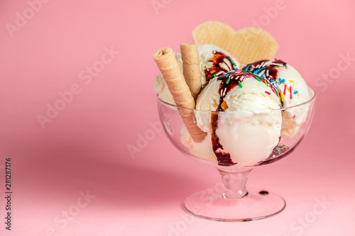 scoops of sundae ice cream in glass bowl with chocolate sauce, strewed sprinkles and cookies on pink background photo