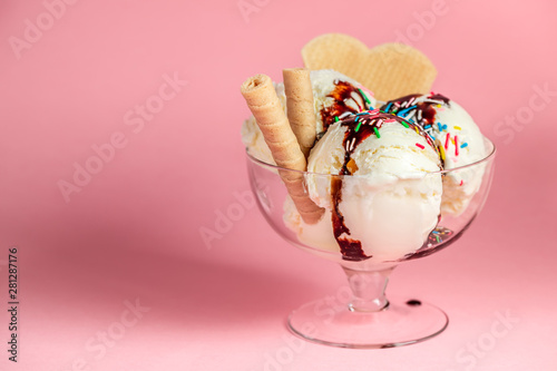 scoops of sundae ice cream in glass bowl with chocolate sauce, strewed sprinkles and cookies on pink background