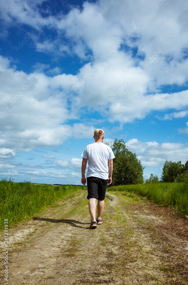 Man walks along a path near a forest in dark shorts and a white T-shirt. Caucasian man with a short haircut back to the camera. Around trees, plants. Place for text. The concept of solitude in nature.