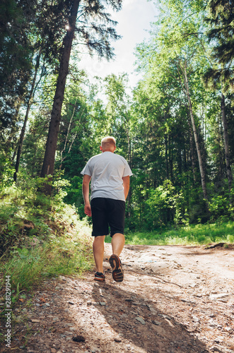 Man walks along a path near a forest in dark shorts and a white T-shirt. Caucasian man with a short haircut back to the camera. Around trees, plants. Place for text. The concept of solitude in nature. © Ольга Холявина
