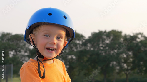 Little boy (kid, child) in orange t-shirt and blue helmet is riding scooter. Childhood memories, safe and funny experience .Closeup portrait, summer, sunny © alenamozhjer