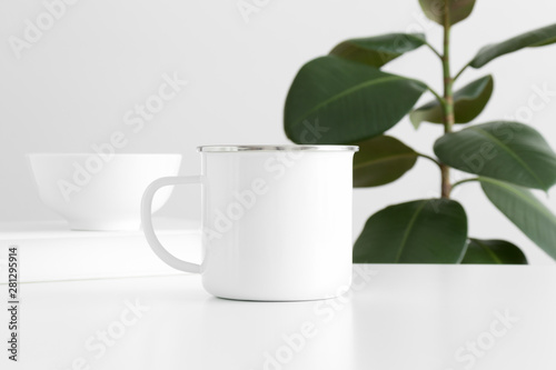 Enamel mug mockup with workspace accessories on a white table and a ficus plant.